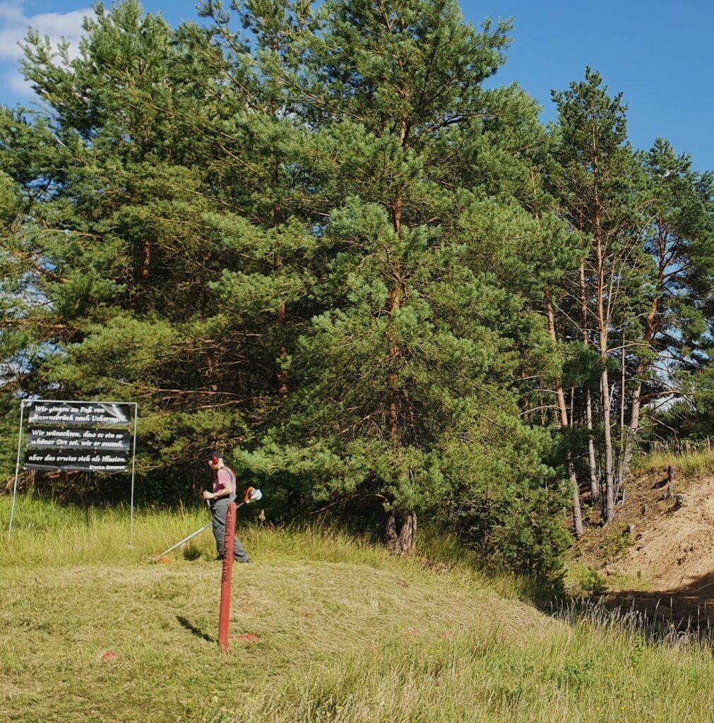 Bildausschnitt vom ehemaligen Lagereingang, zu sehen ist das Transpi mit dem zitat einer Überlebenden, davor senst eine Person Rasen.